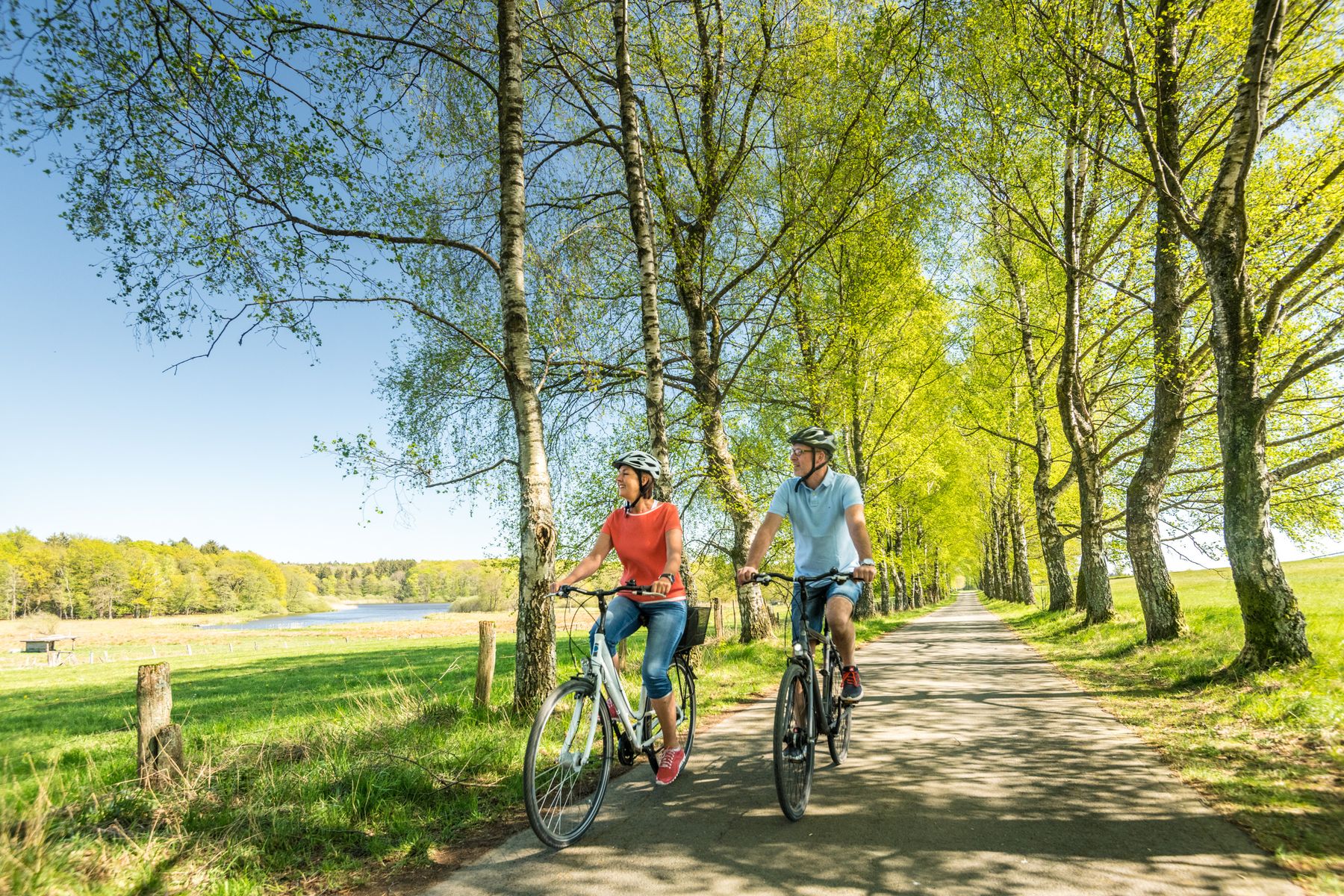 Zwei Personen fahren an einem sonnigen Tag auf einem von Bäumen gesäumten Weg, umgeben von üppigem grünen Gras und Bäumen, Fahrrad. Beide tragen Helme und Freizeitkleidung und genießen die malerische Umgebung in der Nähe eines Gewässers.